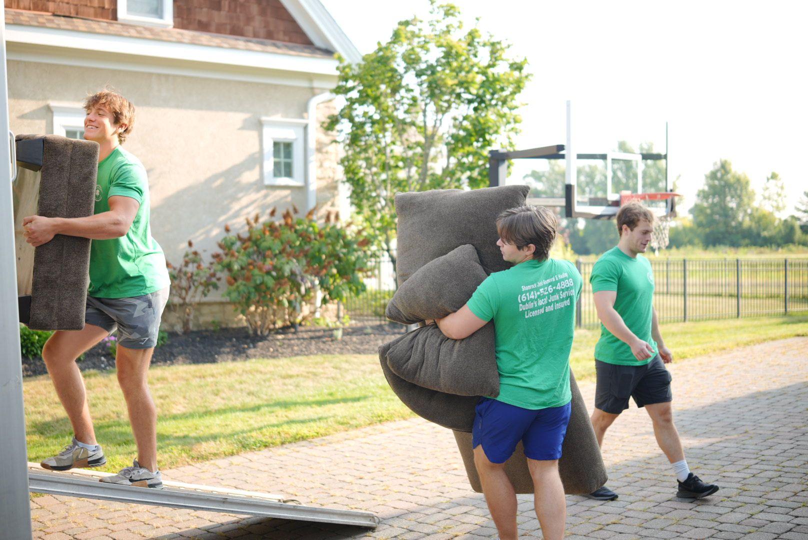 Three Shamrock Junk removal Employees carrying furniture items outdoors near a house and basketball hoop.