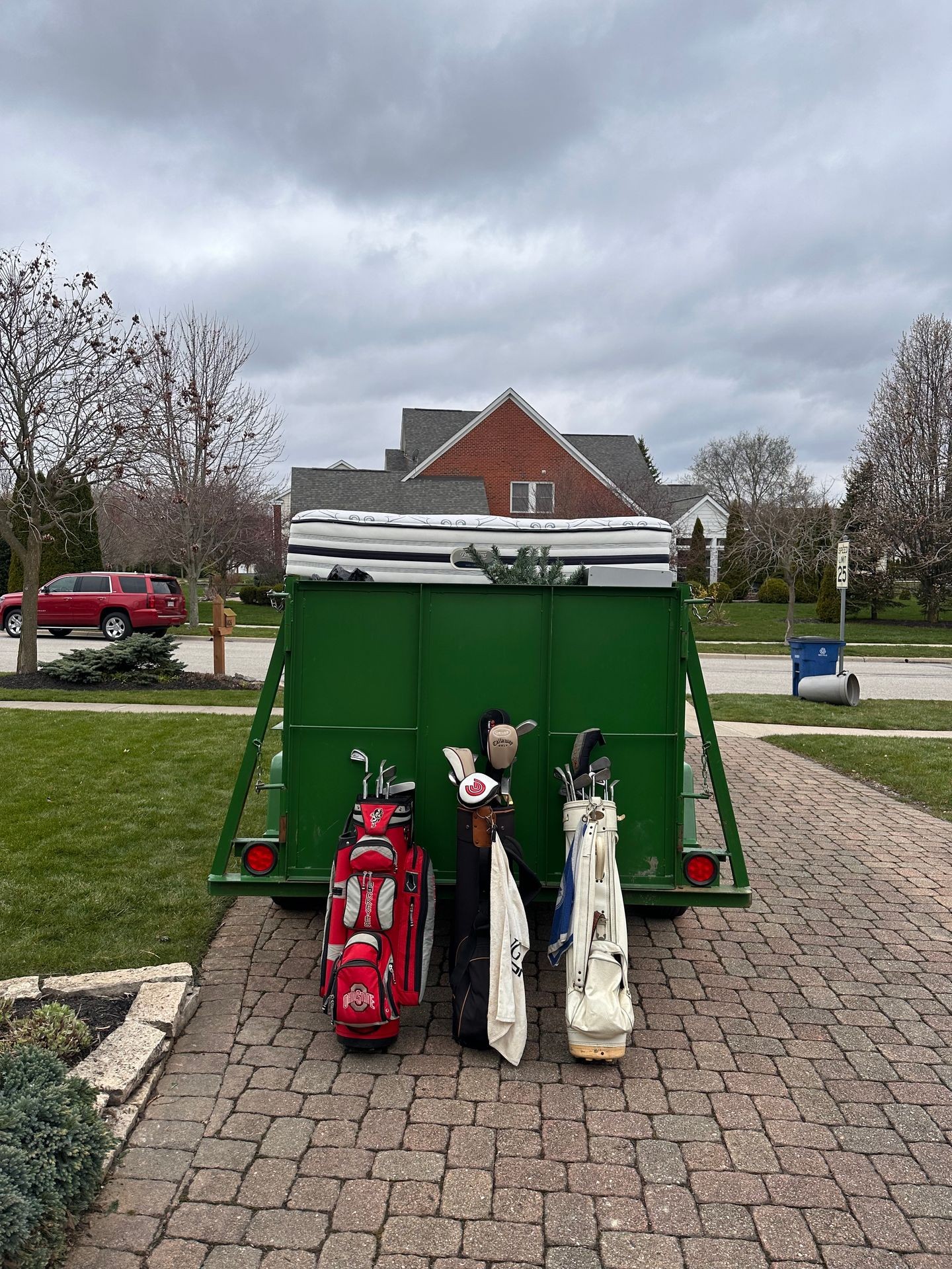 Shamrock Junk Removal removes Golf bags and clubs placed against a green trailer parked on a brick driveway in a suburban neighborhood.