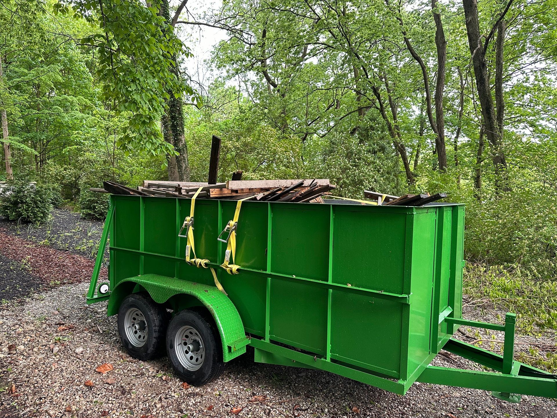 Green Dumpster trailer filled with wooden planks and construction debris, parked on gravel in a wooded area.