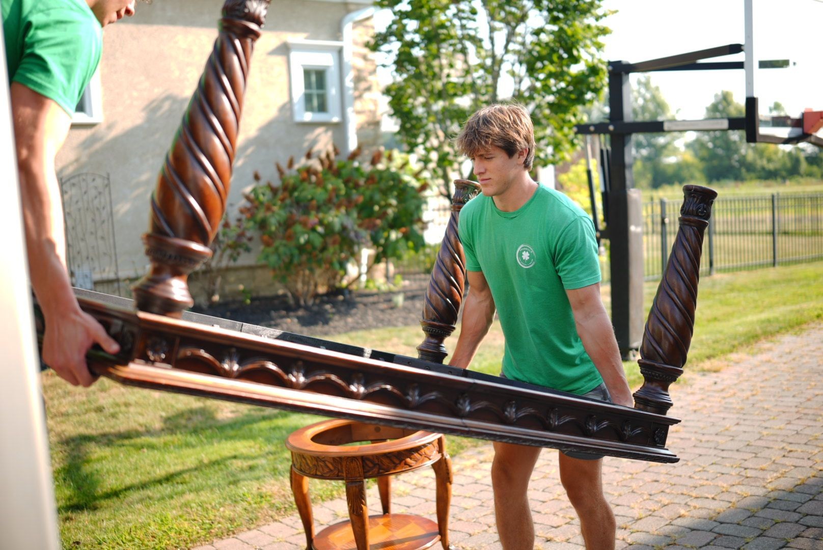 Shamrock Junk Removal employees carry a wooden table with intricate carvings outdoors near a house.